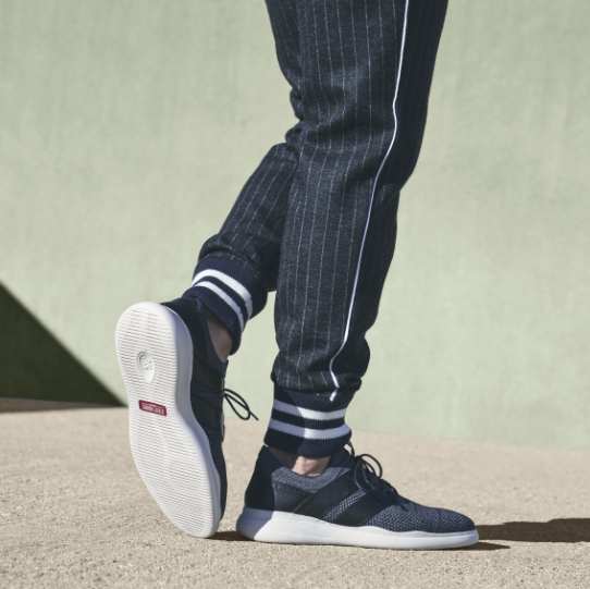 "Work From Home in Style" The featured image is of a model wearing the Moxley Knit Lace Up Sneaker in Black/Grays and Black/White striped casual pants while standing on a sand colored floor.