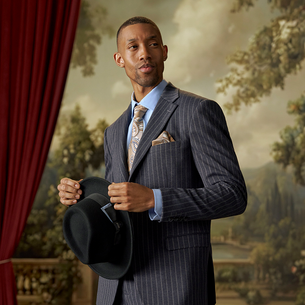 "What To Wear For A Wedding: Outfits For Men." The featured image is of a man in a Stacy Adams dark gray pinstripe suit and light blue shirt holding a black fedora standing against a red curtain and a balcony with green trees.