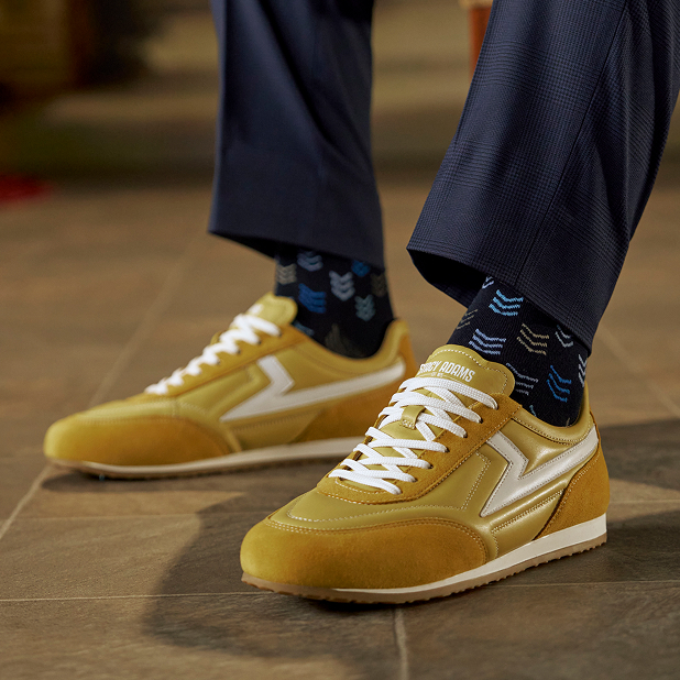 "In The World Of Men’s Casual Shoes, Sneakers Take Center Stage." The featured image is of a model wearing the Action 150 Sporty Lace Ups in yellow with the Mini Chevron Men's Crew Dress Socks in Navy, standing on light brown tile..