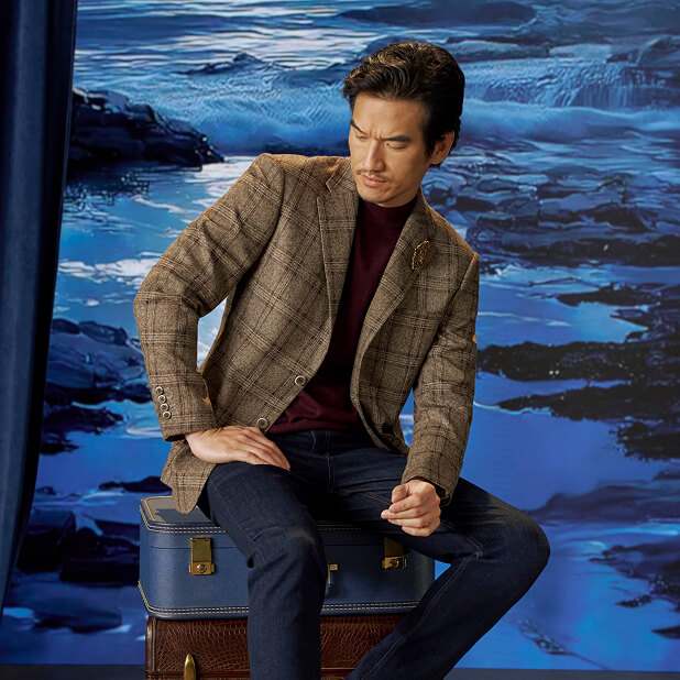 "A Guide To Business Casual Footwear." The featured image is of a model wearing a business casual outfit of a light brown plaid suit jacket over a burgandy shirt with dark wash straight cut jeans, sitting on a stack of two suitcases, one brown and one blue in front of a blue background.