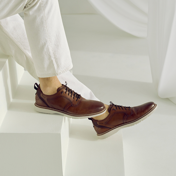 "Sneakers, Loafers, and Casual Shoes For Men That Add Style to Your Downtime." The featured image is of a model wearing the Stacy Adams Sync Plain Toe Elastic Lace Ups in Cognac while sitting on white steps with his legs stretched out.