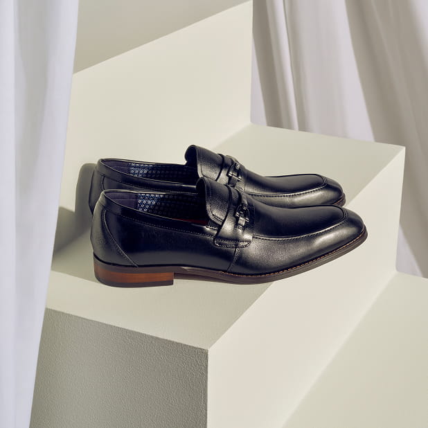 "Why Men’s Black Shoes Are A Wardrobe Must-Have." The featured image shows the Stacy Adams Kaylor Moc Toe Bit Slip Ons in Black sitting on a set of stairs surrounded by white curtains.