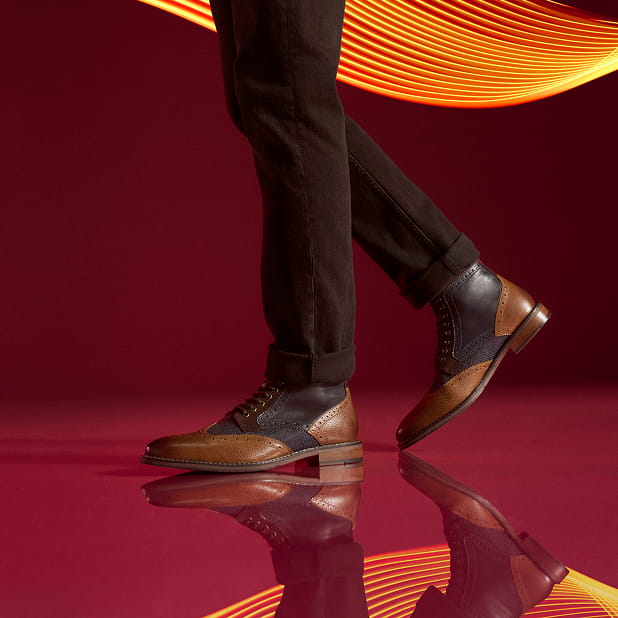 "Men’s Boots: A Storied History." The featured image shows a model walking in the Stacy Adams Finnegan Wingtip Lace Up Boots in Cognac with Navy against a burgandy background with neon lights.