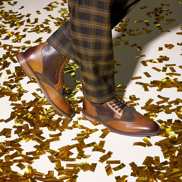 "Men's Boots Throughout The Years." The featured image shows a close-up of a model wearing the Stacy Adams Finnegan Wingtip Lace Up Boots in Olive Multi surrounded by gold confetti on a white background.