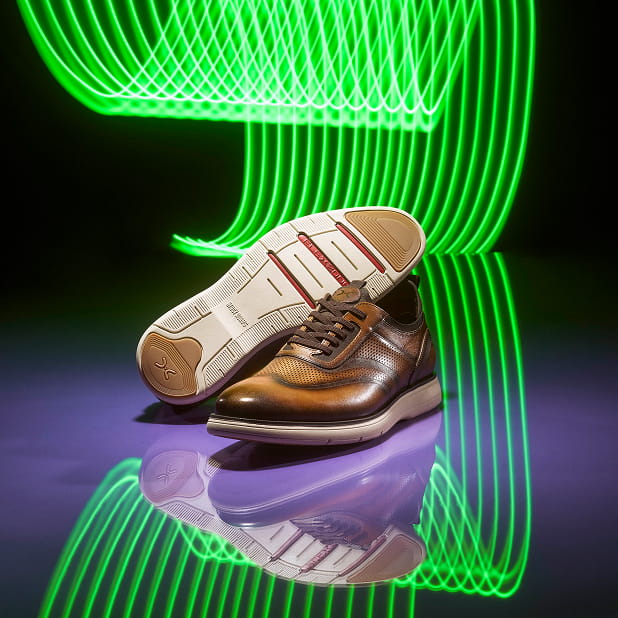 "Men's Travel Tips, The Right Shoes For The Road." The featured image shows the Stacy Adams Edgewood Wingtip Elastic Lace Up in Cognac against a purple background with green neon lights.