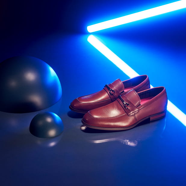 "Tassel, Bit, Or Penny Loafer: The Options Are Endless." The featured image shows the Stacy Adams Kaylor Moc Toe Bit Slip Ons in Cognac on a dark blue background with a neon light and decorative half spheres.